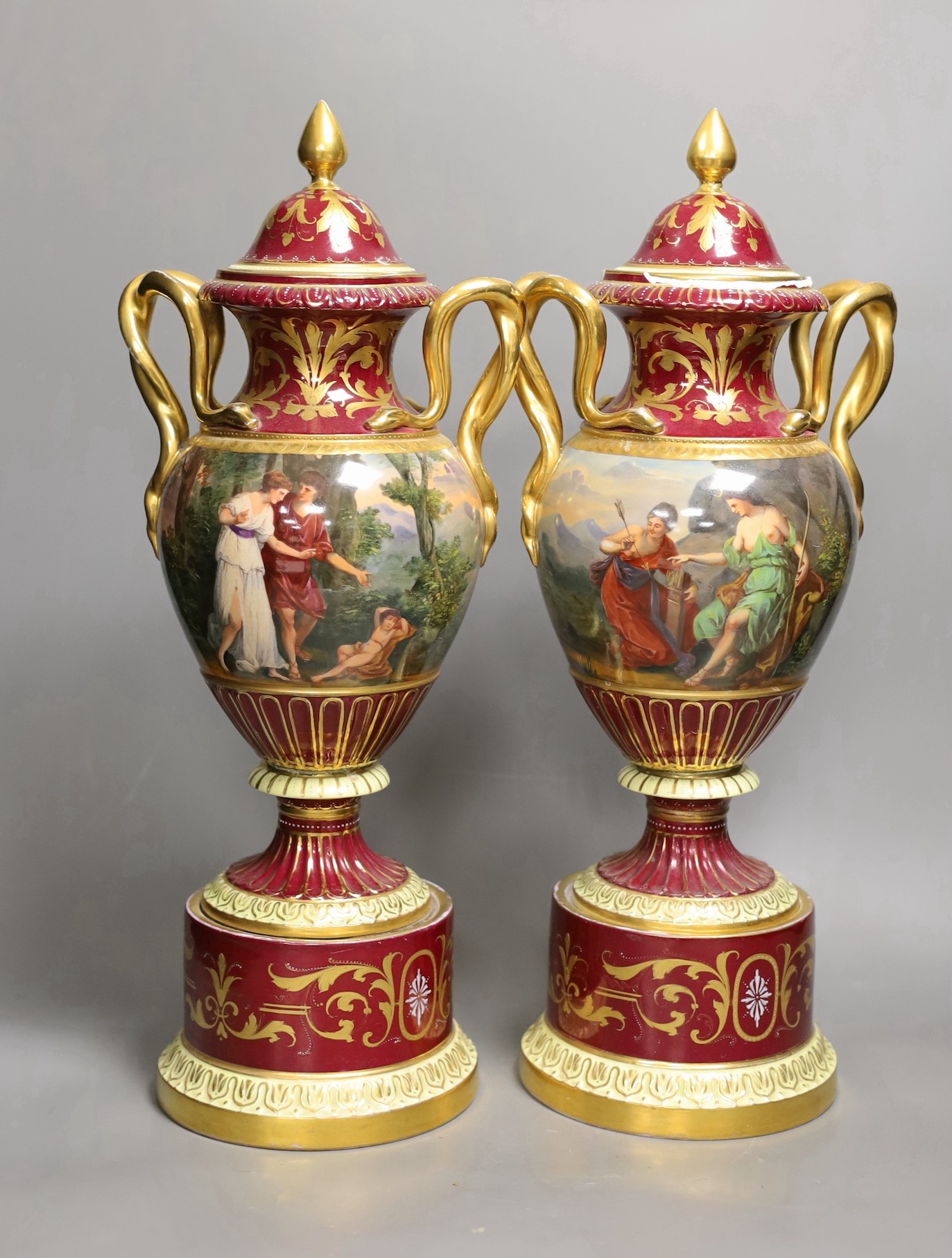 A pair of ornate Vienna style two handled urns bearing covers, with painted love scene and red ground and gilt decoration, 46.5cm high